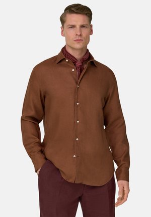 Hombre con camisa marrón de manga larga abotonada y pantalones burdeos, con un pañuelo para el cuello a juego, de pie frente a un fondo liso.