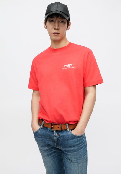 T-shirt en coton rouge avec un motif de poisson blanc et le texte "FERMÉ POUR L'ÉTÉ." Associé à un jean en denim bleu clair et une ceinture en cuir marron.
