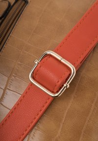 Simili cuir beige avec un motif crocodile, doté d'une sangle orange et d'une boucle métallique de couleur dorée.