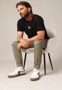 Zwarte t-shirt met korte mouwen en logo, olijfgroene cargo broek, witte sneakers met zwarte strepen, zittend op een gestructureerde crèmekleurige stoel.