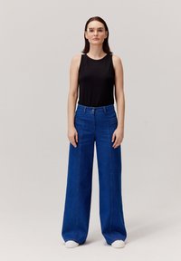 Czarny prążkowany top na rami ączkach i niebieskie spodnie z szerokimi nogawkami. Okulary, białe trampki. Prosty design z zapięciem na guziki i przednimi kieszeniami. Gładka faktura.