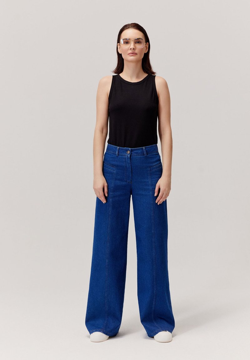 Czarny prążkowany top na ramiączkach i niebieskie spodnie z szerokimi nogawkami. Okulary, białe trampki. Prosty design z zapięciem na guziki i przednimi kieszeniami. Gładka faktura.