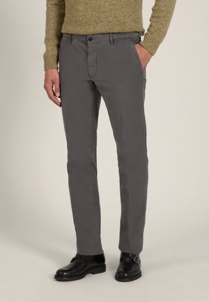 Mann trägt graue Slim-Fit-Hose, schwarze Lederschuhe und einen grünen Strickpullover, steht mit einer Hand in der Tasche vor neutralem Hintergrund.