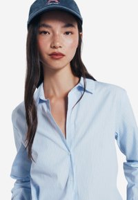 Camicia azzurra chiara con sottili righe verticali, tessuto morbido e colletto; abbinata a un berretto navy con logo.