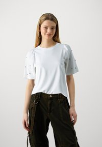 Weißes T-Shirt mit kurzen Puffärmeln und kleinen dekorativen Akzenten. Kombiniert mit dunkelgrünen Cargo-Hosen, die das funktionale Design betonen.