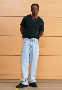 Zwart T-shirt met korte mouwen, lichtblauwe wijde jeans en zwarte schoenen. Eenvoudig ontwerp met een subtiel logo op de tailleband. Effen achtergrond.