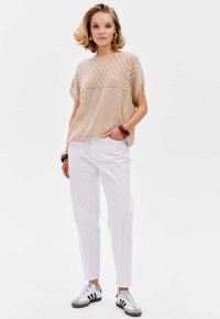 Beżowy dzianinowy top w diamentowy wzór z krótkimi rękawami, w połączeniu z białymi spodniami i szaro-brązowymi trampkami w czarne paski.