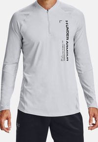 Mies, joka pukeutunut vaaleanharmaaseen pitkähiharaiseen Under Armour -paitaan, jossa on neljännesvetoketju ja musta tekstitunnus edessä, yhdistettynä mustiin shortseihin.