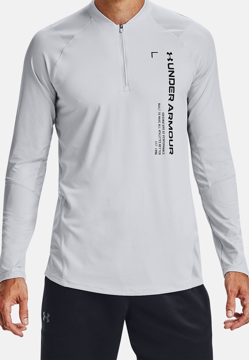 Mies, joka pukeutunut vaaleanharmaaseen pitkähiharaiseen Under Armour -paitaan, jossa on neljännesvetoketju ja musta tekstitunnus edessä, yhdistettynä mustiin shortseihin.