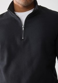 Zwarte zip-up trui met een hoge kraag, voorzien van een zilveren rits en een zachte, gladde textuur, gedragen over een wit shirt.