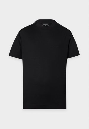 Mann trägt ein schwarzes kurzärmeliges T-Shirt, hellblaue Jeans mit weitem Bein und weiße Turnschuhe, steht mit einer Hand in der Tasche.