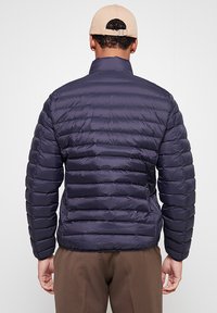 Veste matelassée bleu marine avec coutures horizontales, col montant et design léger. Portée avec un pantalon marron et une casquette beige.