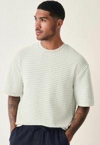 White textured short-sleeve shirt with a round neck, featuring a woven pattern. The model wears navy shorts and has visible tattoos.