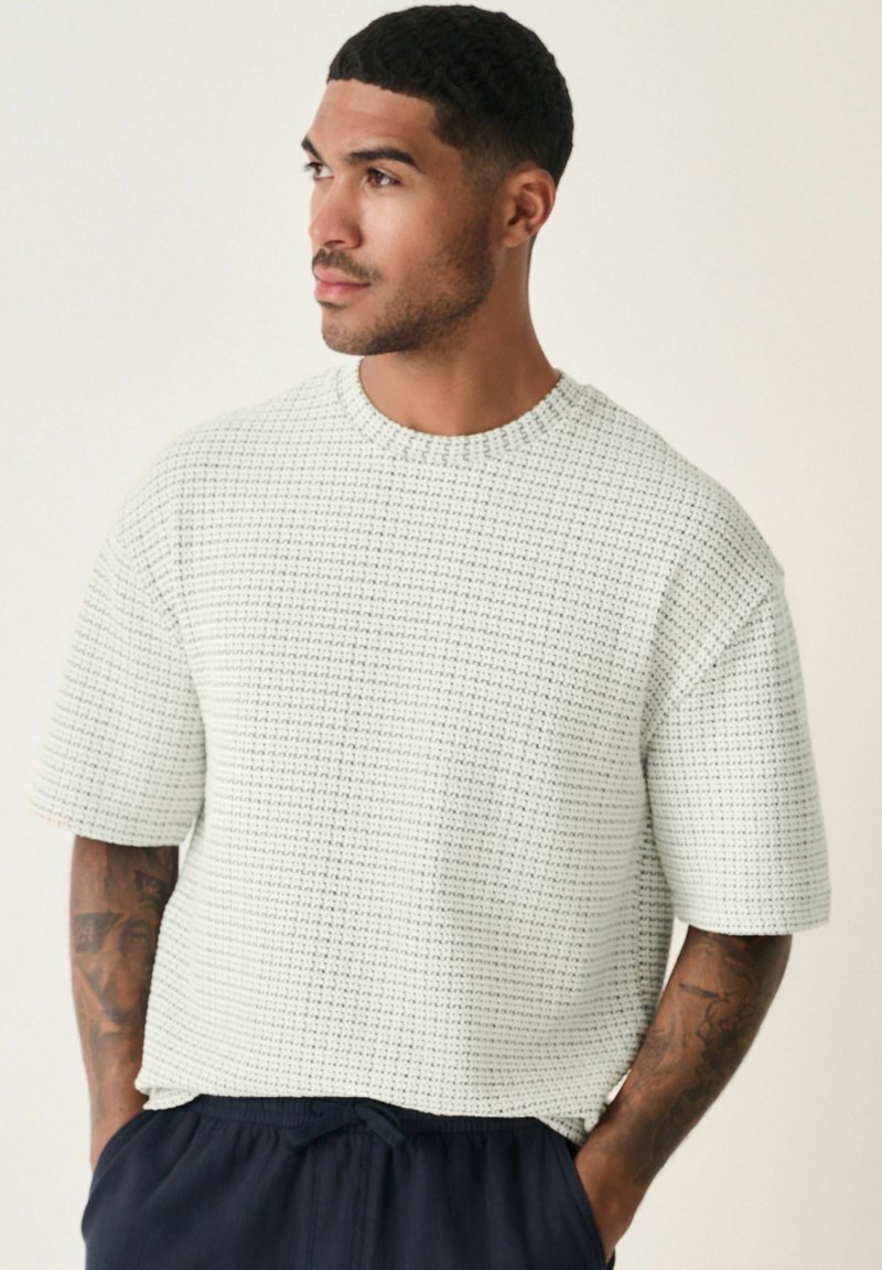 Weißes, strukturiertes Kurzarmhemd mit Rundhalsausschnitt und gewebtem Muster. Das Modell trägt eine marineblaue Shorts und hat sichtbare Tattoos.