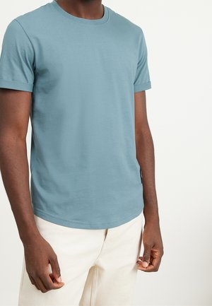 Mann trägt ein hellblaues kurzärmeliges T-Shirt und cremeweiße Hosen vor einem schlichten hellen Hintergrund.