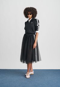 Femme aux cheveux bouclés portant une veste noire Adidas et une jupe en résille à rayures blanches, des lunettes de soleil noires et des ballerines rose pâle.