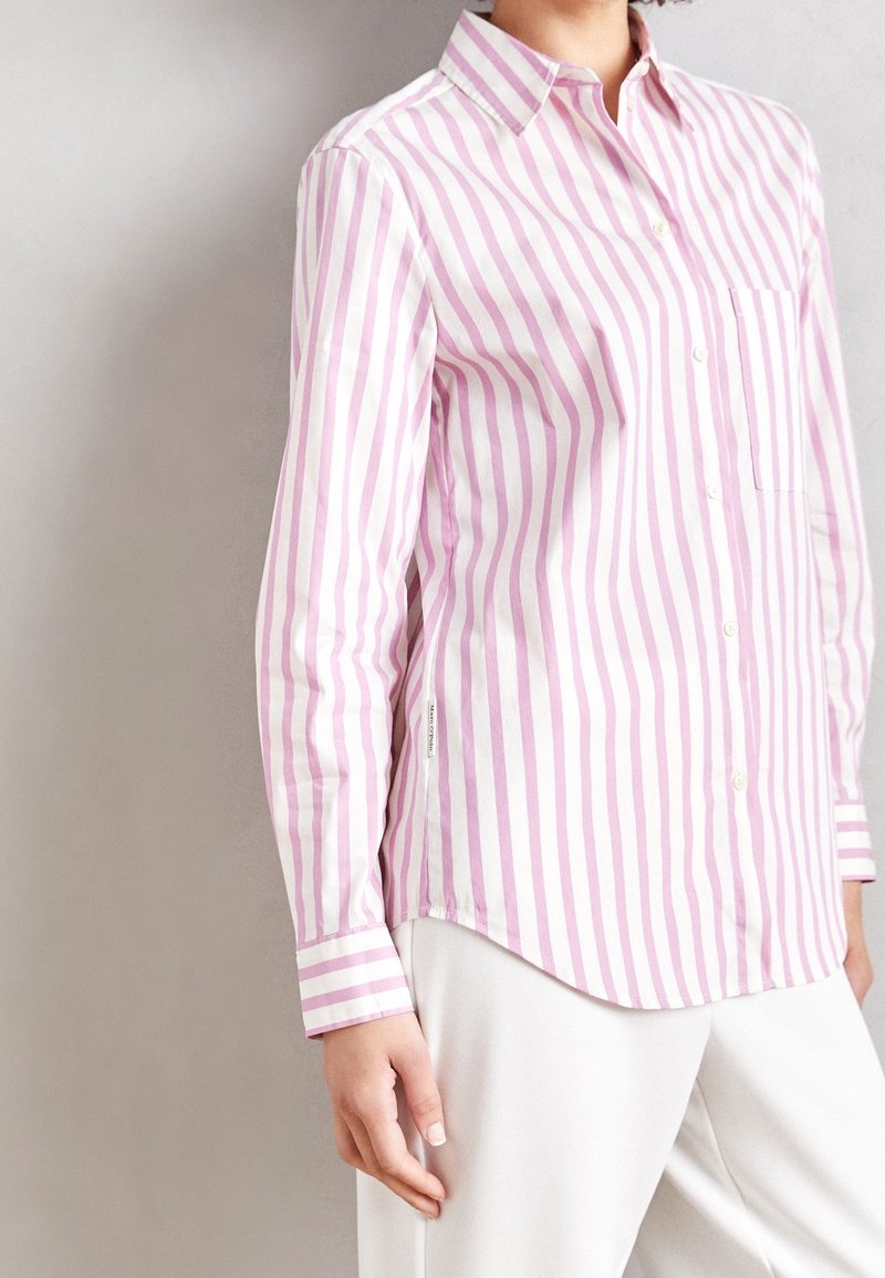 Personne portant une chemise à manches longues boutonnée avec des rayures verticales roses et blanches et un pantalon blanc, debout devant un fond uni.