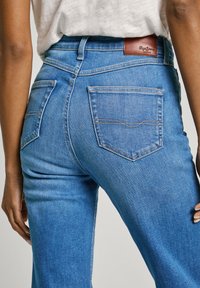 Jean en denim bleu avec une taille mi-haute, deux poches arrière et un patch en cuir marron au niveau de la taille, présentant une texture lisse.