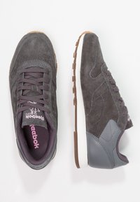 Baskets en suède gris foncé avec semelles en caoutchouc, arborant une touche de violet sur la languette et le talon, ainsi qu'un design texturé sur le dessus.