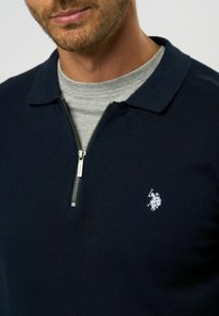 Mornarska zip-up polo majica z teksturirano tkanino, z sivo notranjo ovratnico in vezenim logotipom v beli barvi na levem prsnem delu.