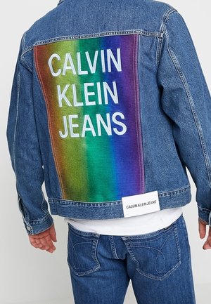 Person trägt blaue Jeansjacke und Jeans, auf dem Rücken der Jacke befindet sich ein großes Regenbogen-Patch mit weißem "Calvin Klein Jeans"-Text.