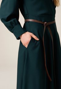 Zelené šaty s dlouhým rukávem z hladké látky, s hnědým koženým opaskem a boční kapsou. Rukáv má knoflíkový detail.