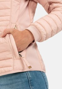 Lyserød jakke med quiltet design, der har en sidelomme og ribbede manchetter. Glat tekstur med kontrastmaterialedetaljer.