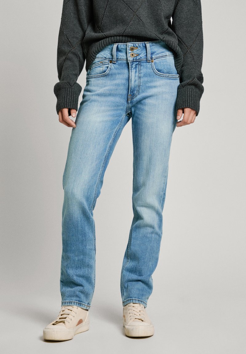 Personne portant un jean droit bleu clair, un pull en maille gris foncé et des baskets beiges à lacets, debout sur un fond gris.