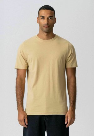 Mand stående med front mod kameraet, iført en ensfarvet beige T-shirt med korte ærmer og mørke bukser mod en lysegrå baggrund.