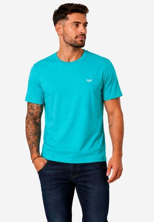 Hombre con barba y tatuajes que lleva una camiseta de manga corta turquesa y vaqueros oscuros, de pie con una mano en el bolsillo, mirando hacia la derecha.