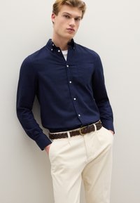 Camisa azul marino abotonada con bolsillo en el pecho, combinada con pantalones beige claro y un cinturón trenzado marrón con hebilla circular dorada.