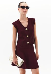 Burgunderfarbene ärmellose Bluse mit drei goldenen Knöpfen und passendem ausgestelltem Rock, kombiniert mit einer goldenen metallischen Clutch und schwarzen Sonnenbrille.