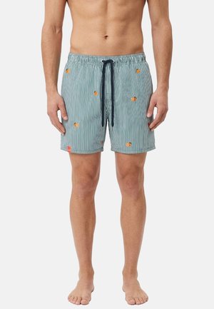 Uomo che indossa pantaloncini da bagno a righe verticali blu e bianche con piccoli motivi di granchi arancioni e un cordino nero, a piedi nudi su sfondo bianco.