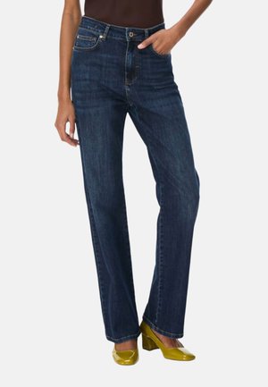 Donna che indossa jeans blu scuro a vita alta con gamba dritta e scarpe gialle con tacco largo, in piedi davanti a uno sfondo semplice.