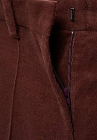 Rjav strukturiran material s žepom na zadrgo; ima maroon zadrgo in pravokotno kovinsko dekoracijo, kar poudarja sodoben dizajn.