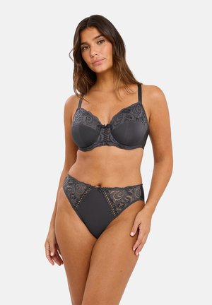 Vrouw die een donkergrijze kanten beha en bijpassende high-waisted slipjes modelleert, staand in een ontspannen houding tegen een effen lichte achtergrond.