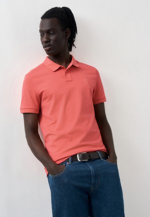 Νέος άνδρας με σκουρόχρωμο δέρμα και dreadlocks, φοράει ένα κοραλλί πόλο και μπλε τζιν, στέκεται με τα χέρια στις τσέπες, κοιτώντας αριστερά.