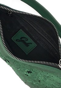 Interno di una borsa a tracolla verde con paillettes, foderato in morbido nero, con una cerniera argentata e un'etichetta in pelle verde con il nome del marchio impresso.