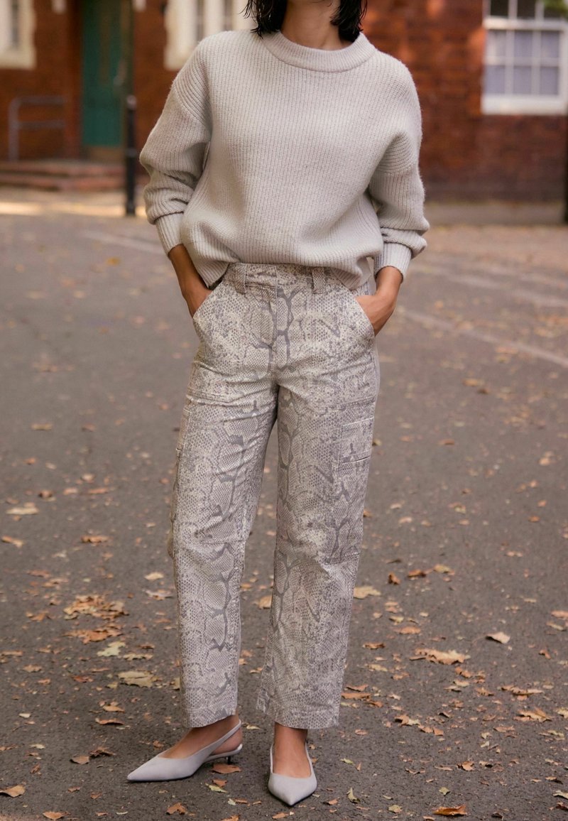 Femme portant un pull beige ample, un pantalon taille haute à motif serpent, et des chaussures slingback pointues gris clair, debout dans une rue bordée de feuilles.