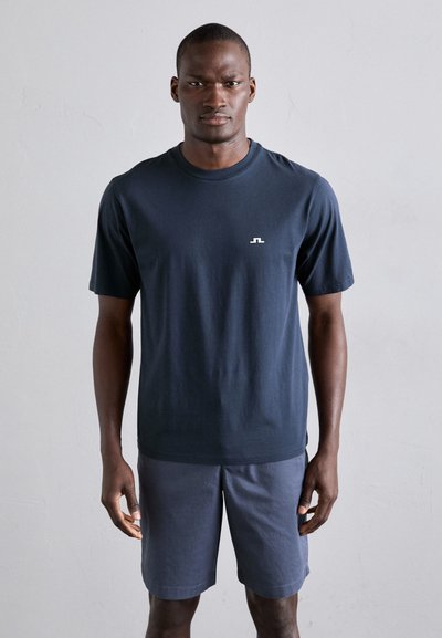 Marineblauw katoenen T-shirt met korte mouwen en een ronde hals, voorzien van een klein wit logo op de borst. Gecombineerd met grijze shorts.