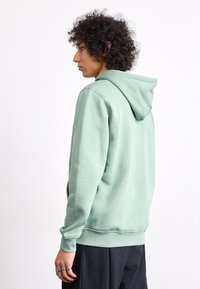 Sweatshirt verde claro com uma textura suave, capuz com cordão e punhos canelados. O design apresenta um corte confortável e bolsos tipo canguru.