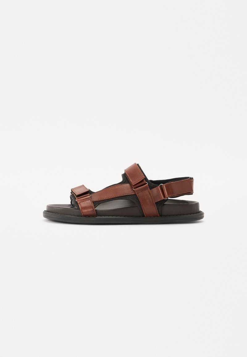 Sandalias de cuero marrón con correas ajustables, suela plana y un diseño minimalista. Presenta punta y talón abiertos con detalles en negro.