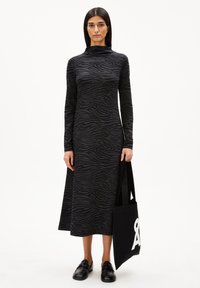 Frau in einem schwarzen, zebramusterigen Midi-Kleid mit langen Ärmeln, die eine schwarze Tote-Bag mit weißem Text hält, trägt schwarze Slipper und steht vor einem weißen Hintergrund.