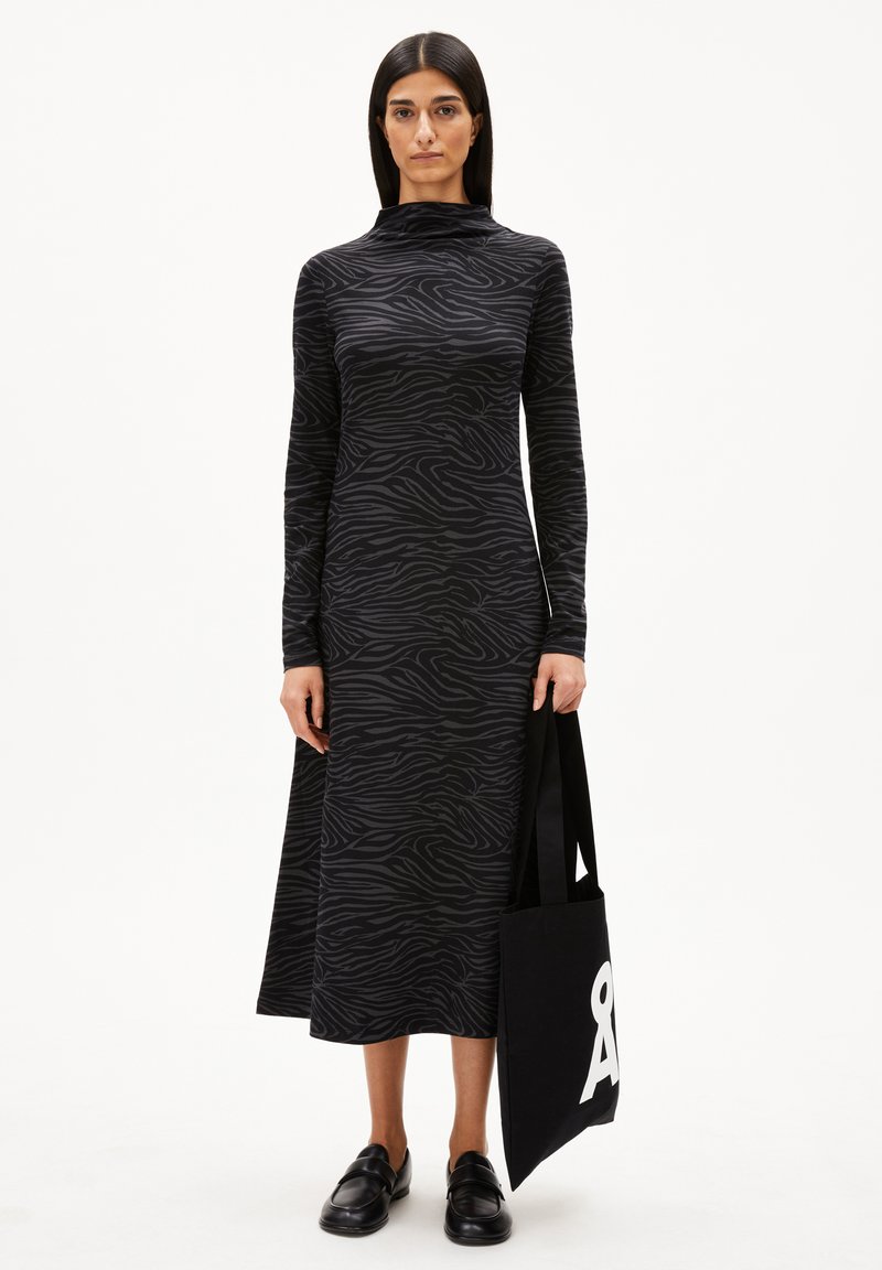 Frau in einem schwarzen, zebramusterigen Midi-Kleid mit langen Ärmeln, die eine schwarze Tote-Bag mit weißem Text hält, trägt schwarze Slipper und steht vor einem weißen Hintergrund.