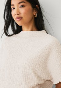 Hellbeige strukturierte Bluse mit hohem Kragen und weiten Ärmeln. Das Model trägt goldene Creolen und hat lange, glatte Haare.