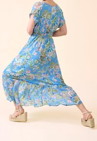 Robe maxi à fleurs bleu clair avec un design fluide, des manches courtes et un bas à volants. Portée avec une sandale compensée montrant une bride texturée.