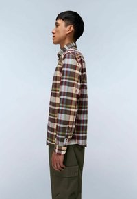 Uomo in piedi di lato che indossa una camicia a quadri multicolore e pantaloni cargo verde oliva su uno sfondo chiaro e semplice.