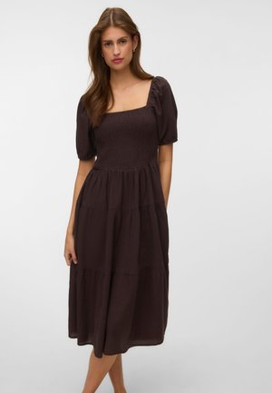 Femme portant une robe midi marron foncé à volants avec des manches courtes bouffantes et un décolleté carré, debout devant un fond uni clair.
