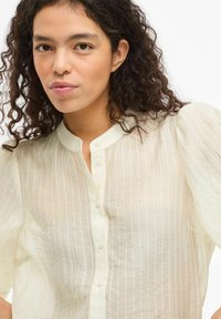 Blusón de crema claro, abotonado, con rayas verticales y sutiles toques brillantes. Cuenta con un collar redondo y mangas abullonadas de tela transparente.