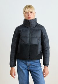 Svart dunjakke med høy krage, som har et mykt, fleece-lignende bunnpanel og elastiske mansjetter. Glidelås foran.